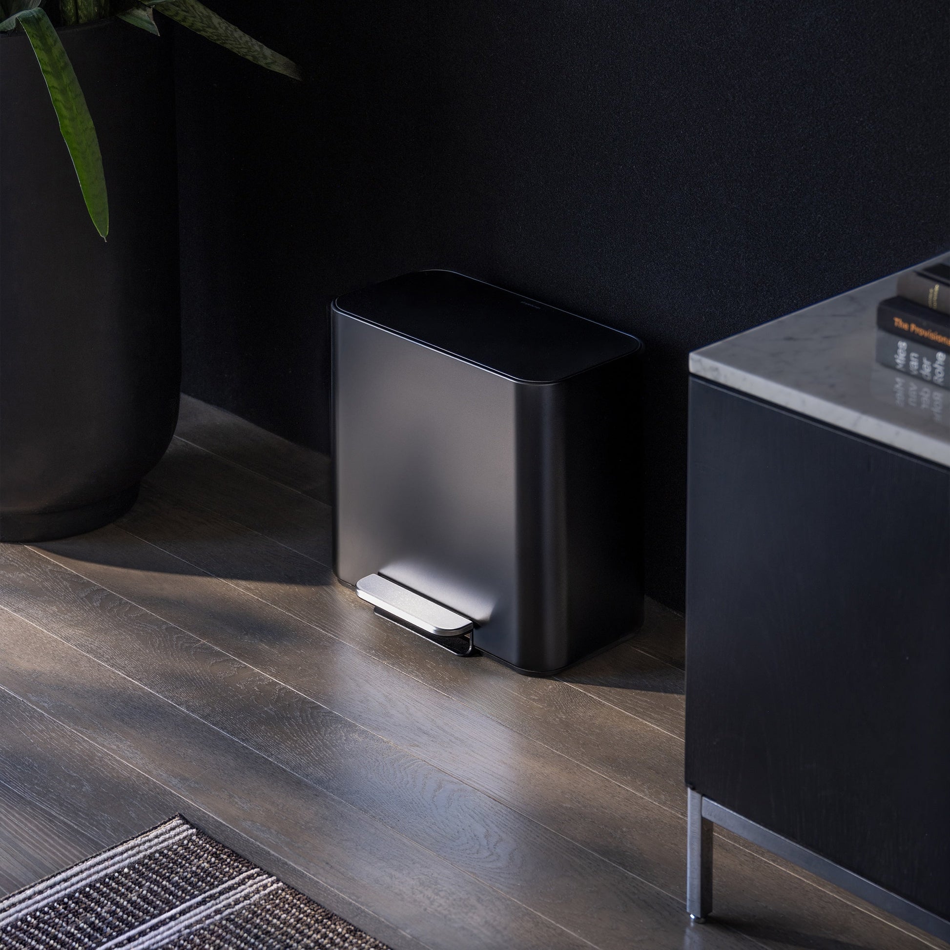 Simplehuman 16L Matte Black Mini Step Can with foot pedal, positioned on wood flooring next to a black storage cabinet with books next to it and plants on the other side.