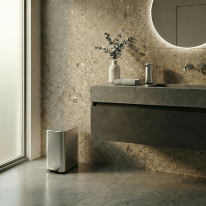 Simplehuman's 5L Slim Step Can in Brushed in a modern bathroom setting. The background also features extra bathroom appliances featuring the Simplehuman Sensor Pump 9 oz. in Brushed along with plants and towels.